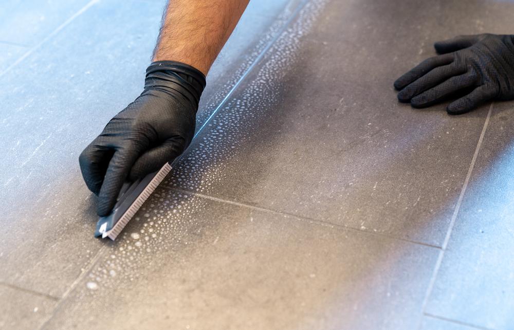 A close up of professional cleaner cleaning grout with a brush blade and foamy soap on a gray tiled bathroom floor